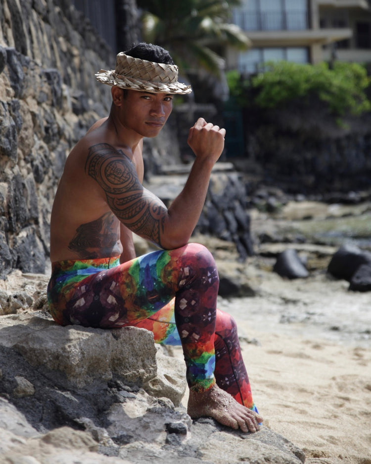 Tattooed man in a woven hat sitting on a beach rock in rainbow crystal-print leggings