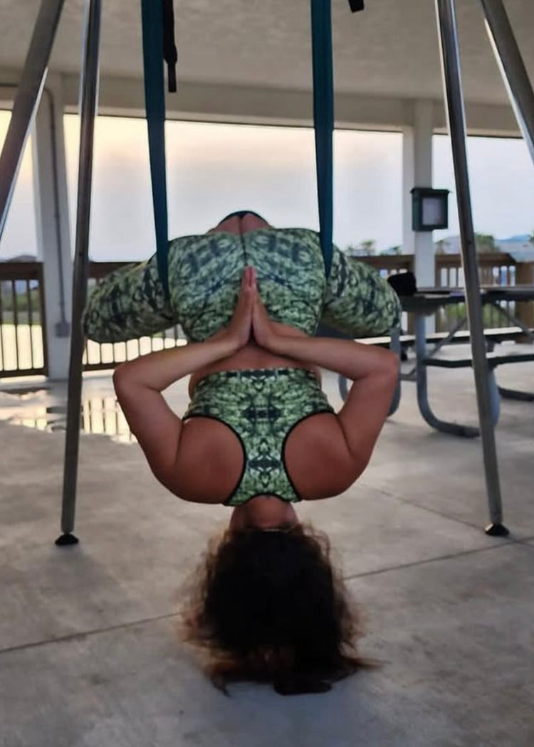 Woman hanging upside down in an aerial yoga hammock in a matching green print sports bra and leggings