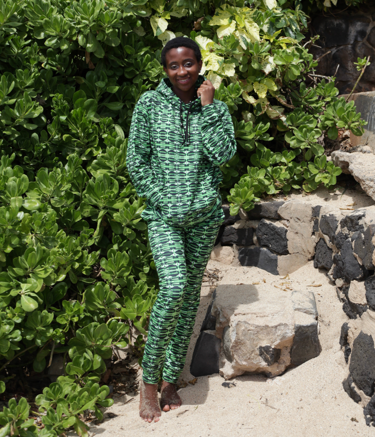 Smiling woman in a matching green print hoodie and joggers standing barefoot on a sandy path beside green shrubs