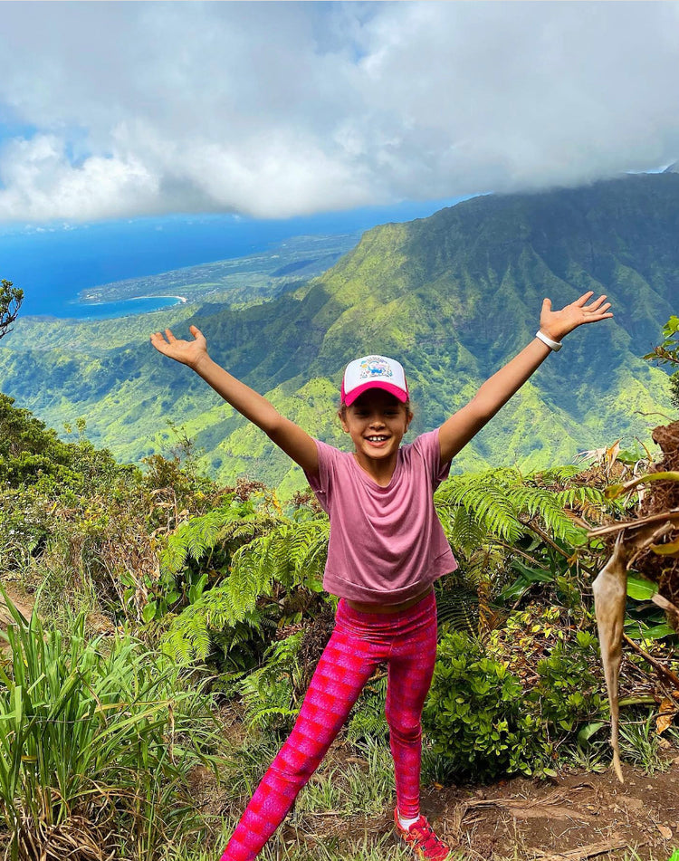 Smiling girl with arms raised on a mountain trail above a green valley and ocean, wearing pink printed leggings