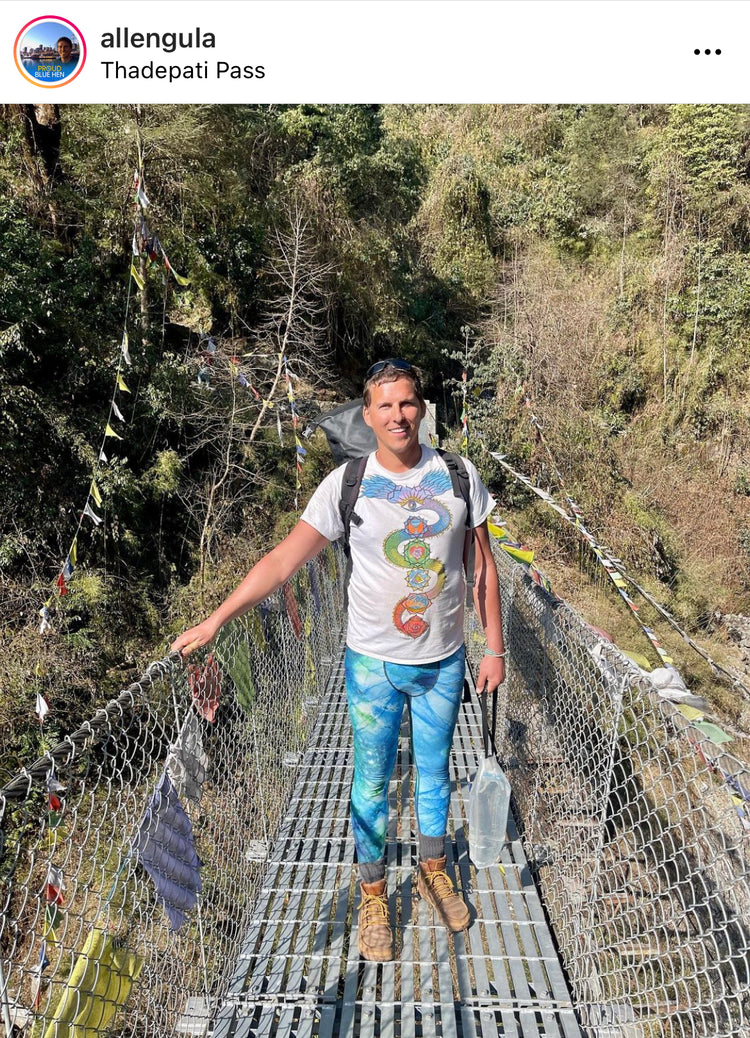 Smiling hiker in blue crystal-print leggings crossing a suspension bridge strung with prayer flags