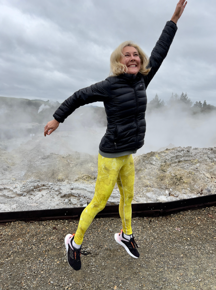 Woman leaping in yellow printed leggings in front of steaming geothermal vents