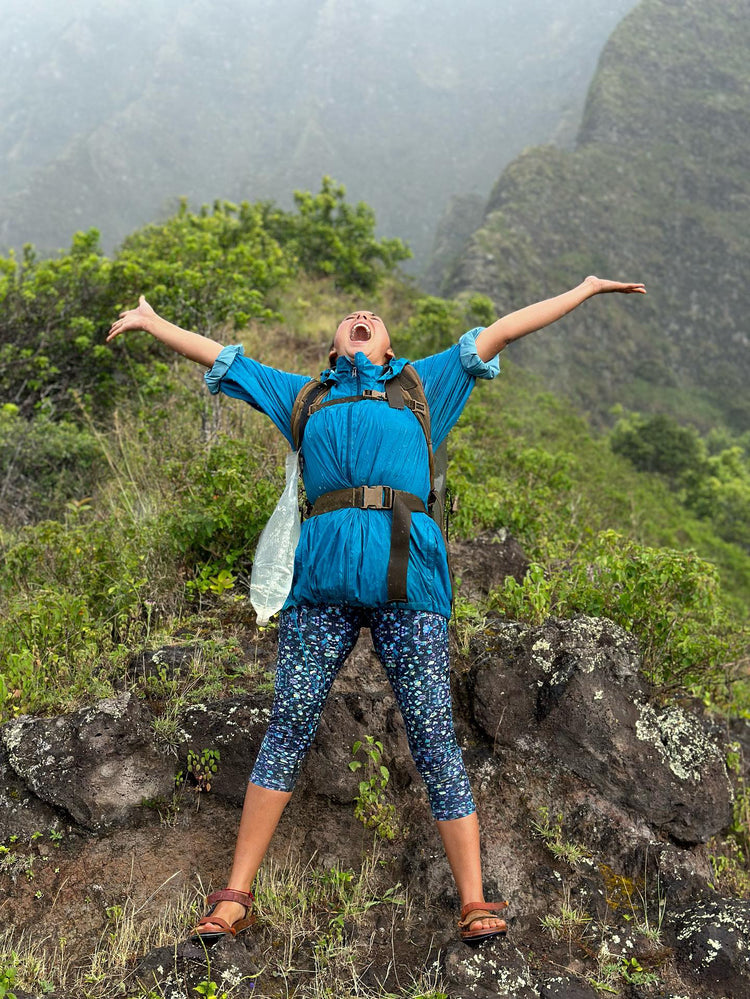 Hiker cheering with arms wide on a misty mountain ridge in blue patterned capri leggings
