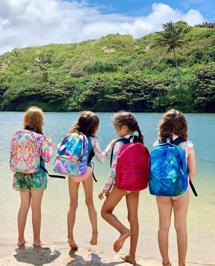 Four girls on a beach facing a turquoise river, each wearing a different colorful crystal-print backpack