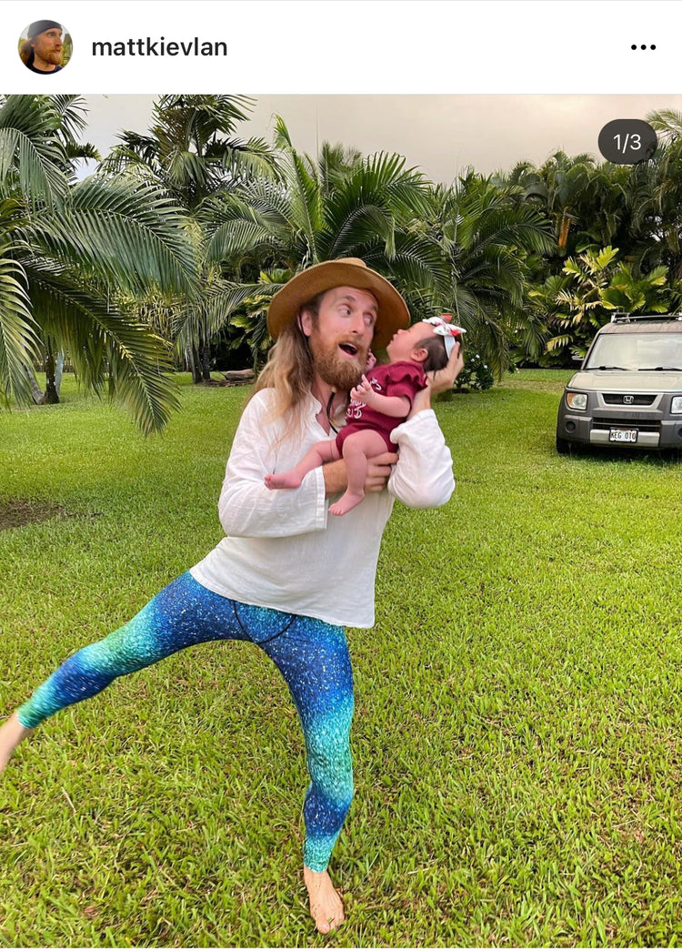Man in a straw hat and blue-green ombre crystal-print leggings dancing on a lawn while holding a laughing baby