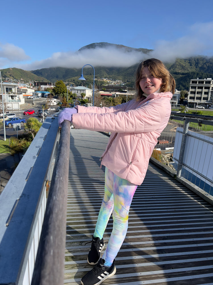 Girl in a pink puffer jacket and pastel tie-dye print leggings on a footbridge with misty green hills behind