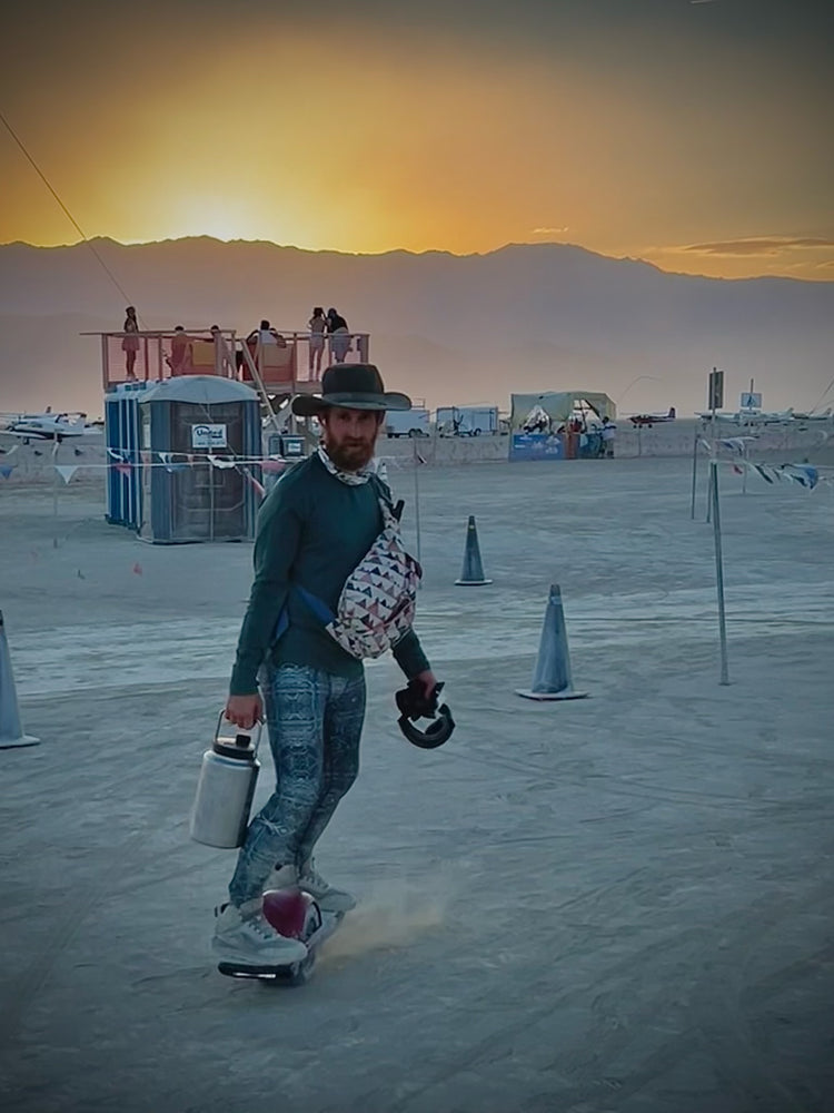 Man in a hat riding a one-wheel board across the desert at sunset in blue print leggings, carrying a water jug