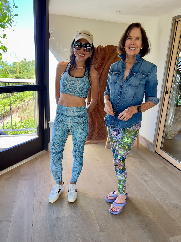 Two smiling women indoors, one in a matching blue feather-print sports bra and leggings, the other in a denim shirt and green and purple crystal-print leggings