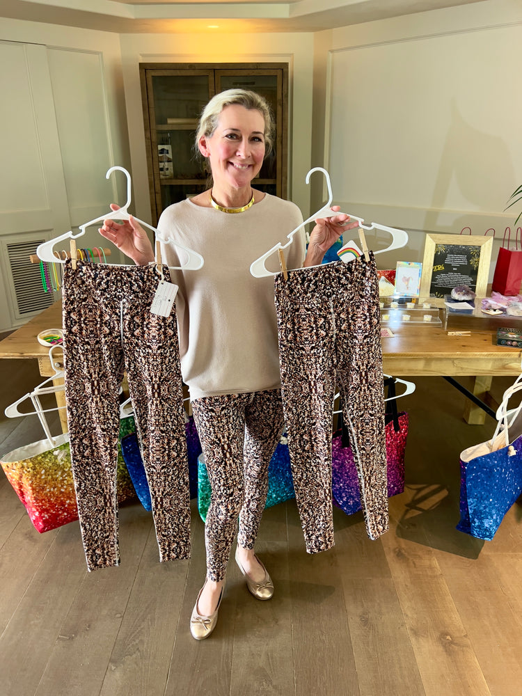 Smiling woman at a pop-up shop holding up two pairs of crystal-print leggings on hangers while wearing a matching pair