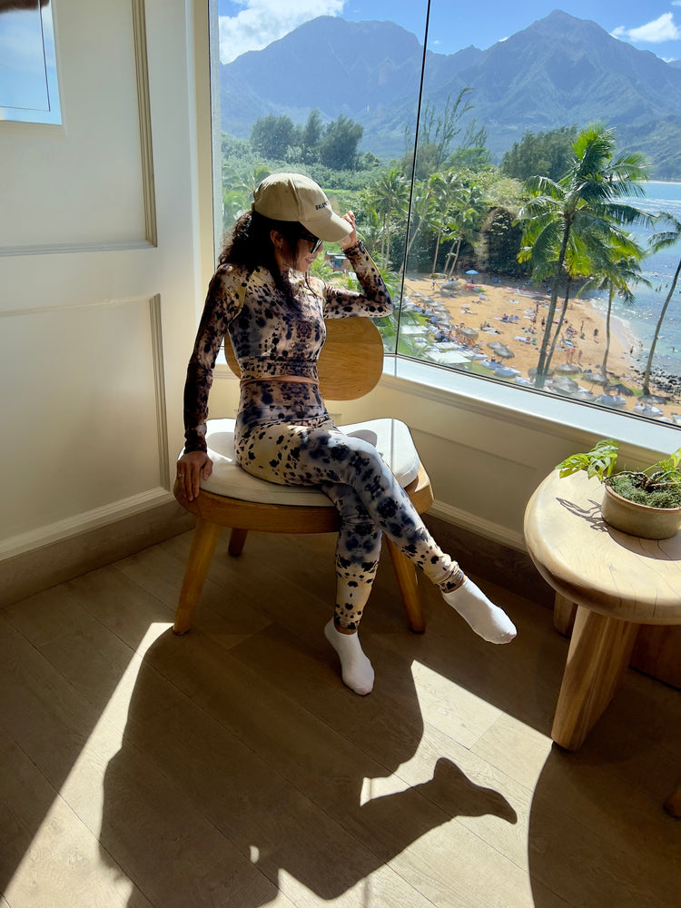 Woman in a cap sitting by a window overlooking a palm-lined bay and mountains, in a matching black and white stone-print long-sleeve top and leggings