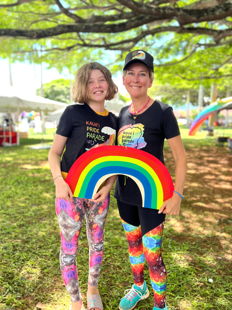 Smiling girl and woman at the Kauaʻi Pride Parade holding a rainbow cutout, both in colorful crystal-print leggings