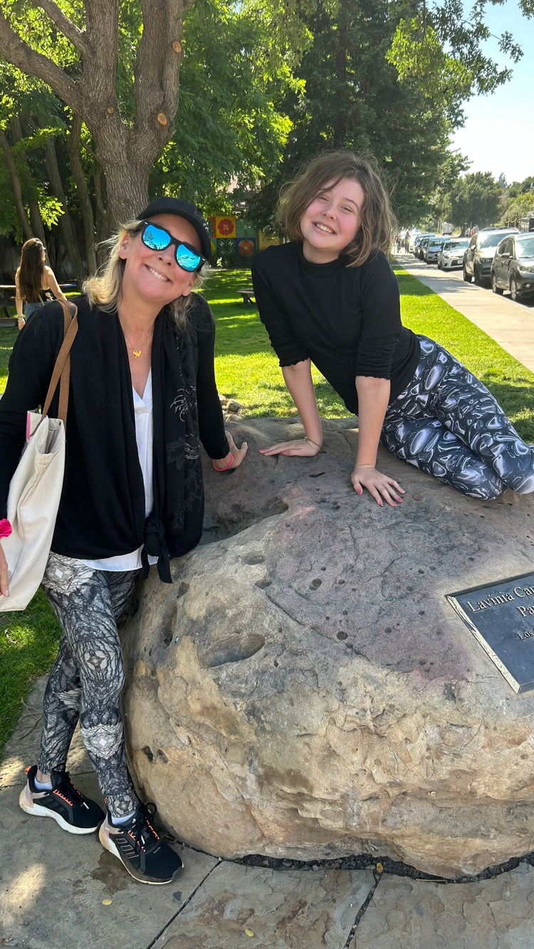 Woman and girl smiling on a large boulder in a park, both wearing patterned leggings