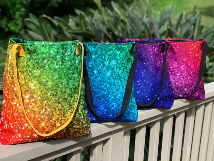 Four sparkling printed tote bags in rainbow colors lined up on a garden railing