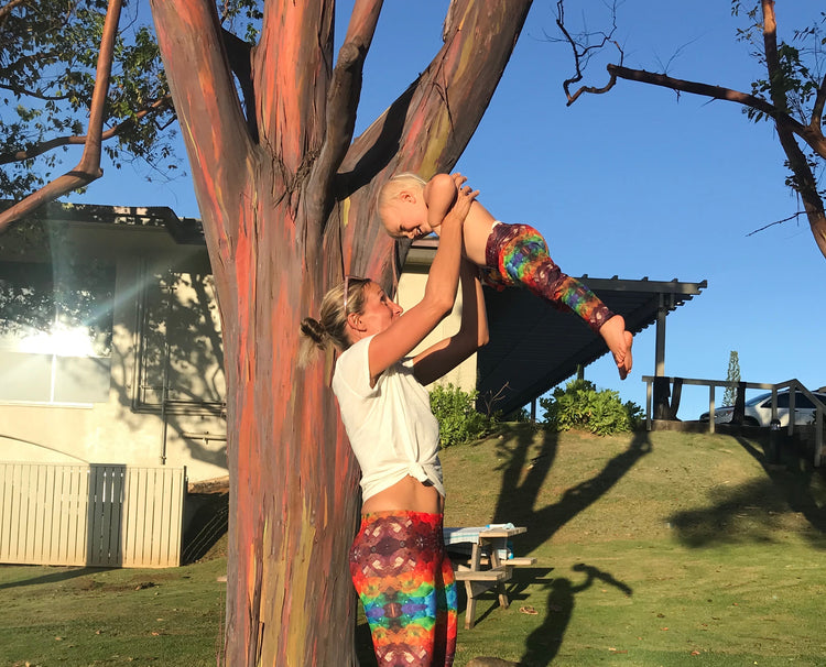 Woman in rainbow crystal-print leggings lifting a toddler in matching leggings beside a rainbow eucalyptus tree